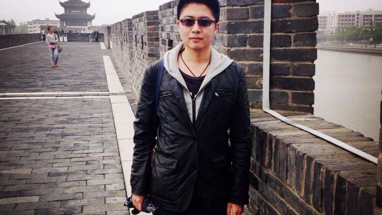 在一个灰色的日子里，一个人戴着太阳镜，穿着轻便的夹克，在砖墙前摆姿势. 背景中可以看到中国传统建筑和现代建筑. 